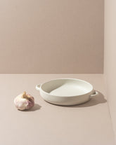 A white stoneware dinner plate with handles, displayed on a neutral background alongside a bulb of garlic.